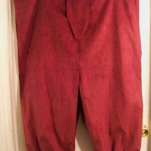 Op de afbeelding: Bordeauxkleurige kniebroek met een knoopsluiting en gerimpelde manchetten. De broek is gemaakt van een zacht, mogelijk su&egrave;de-achtig materiaal en hangt aan een hanger. De stijl suggereert historische of theatrale kleding.