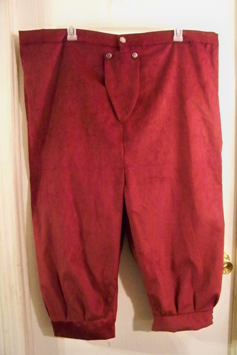 Op de afbeelding: Bordeauxkleurige kniebroek met een knoopsluiting en gerimpelde manchetten. De broek is gemaakt van een zacht, mogelijk su&egrave;de-achtig materiaal en hangt aan een hanger. De stijl suggereert historische of theatrale kleding.