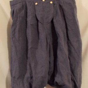 Op de afbeelding: Donkerblauwe linnen bloomers met een hoge taille en geplooide, volumineuze pijpen. De shorts hebben een knoopsluiting en hangen aan een goudkleurige hanger. De stof heeft een licht gekreukte textuur.