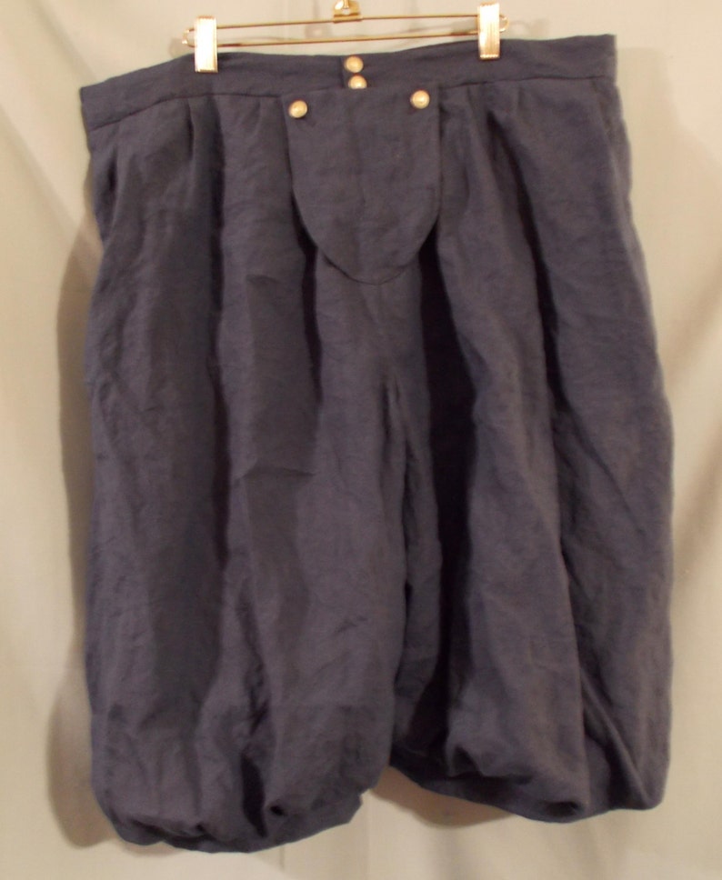 Op de afbeelding: Donkerblauwe linnen bloomers met een hoge taille en geplooide, volumineuze pijpen. De shorts hebben een knoopsluiting en hangen aan een goudkleurige hanger. De stof heeft een licht gekreukte textuur.