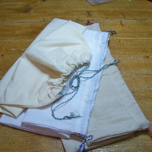 Puede incluir: Una colección de bolsas de tela con cordón en varios tamaños y colores. Las bolsas están hechas de materiales crema, blancos y beige. Cada bolsa tiene un cierre de cordón hecho de cordón trenzado.