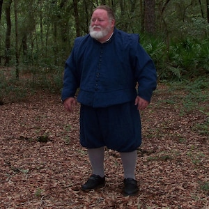 Op de afbeelding: Een persoon in een donkerblauw historisch kostuum, bestaande uit een jas met knopen, knielange broek en zwarte schoenen. De persoon staat in een bos met gevallen bladeren op de grond.