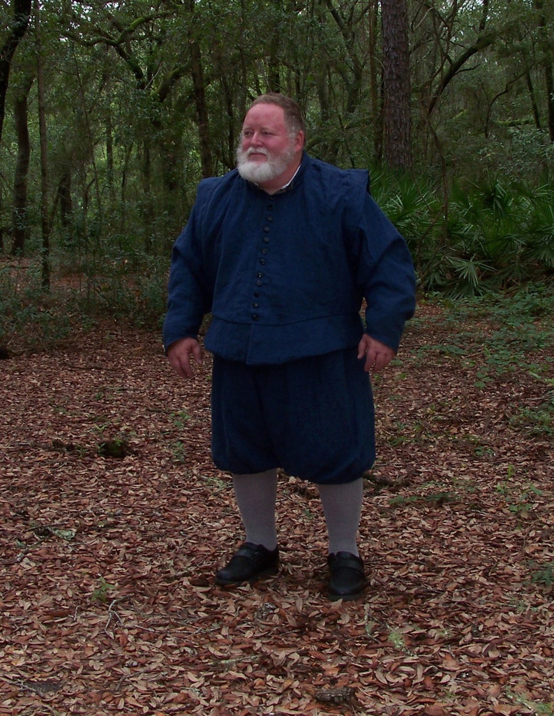 Op de afbeelding: Een persoon in een donkerblauw historisch kostuum, bestaande uit een jas met knopen, knielange broek en zwarte schoenen. De persoon staat in een bos met gevallen bladeren op de grond.