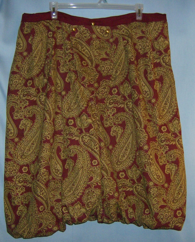 Op de afbeelding: Bordeaux rok met een gouden paisley patroon. De rok heeft een geplooide, ballonachtige vorm aan de onderkant en hangt aan een witte hanger. Gouden knopen sieren de tailleband.