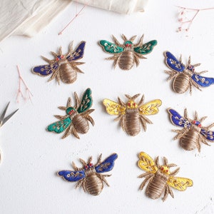 May include: Eight embroidered bee patches in various colours, including blue, green, and yellow. The bees are made of gold thread and have red bead eyes.