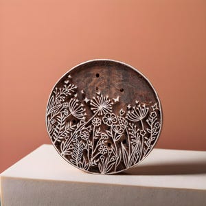 May include: A round wooden block with a floral design carved into the surface. The design features a variety of flowers and leaves, including dandelions, and is carved in a white color against the brown wood.