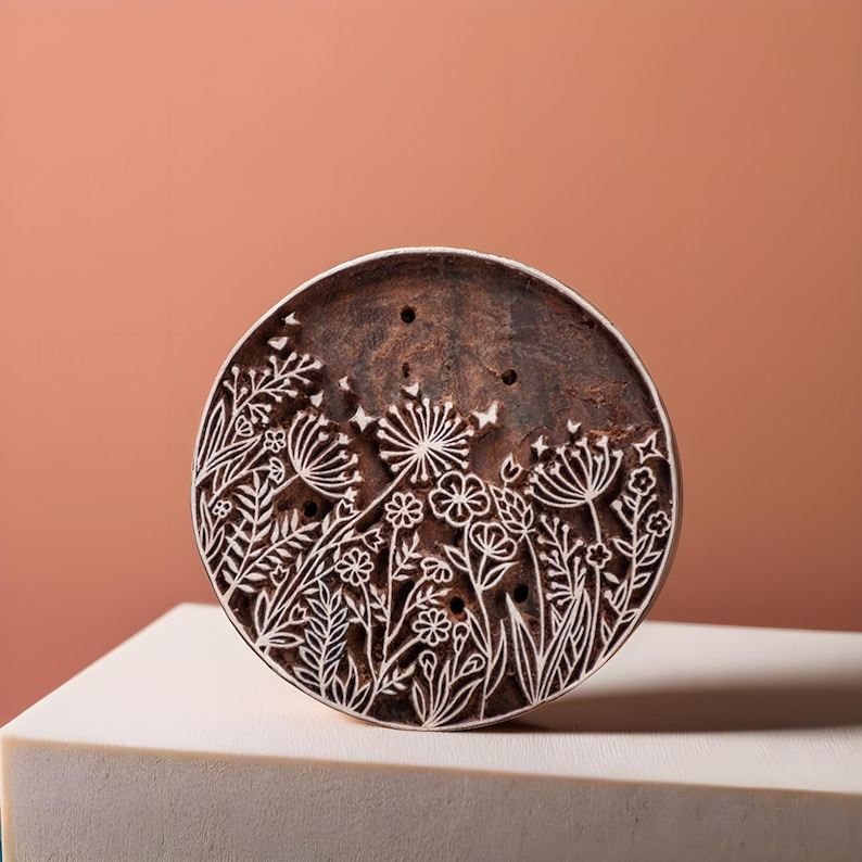 May include: A round wooden block with a floral design carved into the surface. The design features a variety of flowers and leaves, including dandelions, and is carved in a white color against the brown wood.