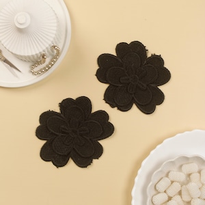 Può includere: Due applicazioni di fiori in tessuto nero con un design ricamato in rilievo. Le applicazioni sono su una superficie beige chiaro.