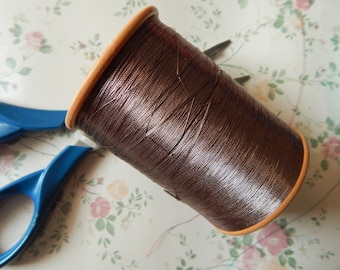 Könnte beinhalten: Eine Spule braunen Fadens mit einem Holzkern. Der Faden ist eng gewickelt und hat einen leichten Glanz. Blaue Scheren sind im Hintergrund teilweise sichtbar, was auf ein Näh- oder Bastelthema hindeutet.