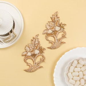 May include: Two gold floral embroidery patches with white beads and sequins. The patches are on a light yellow background.