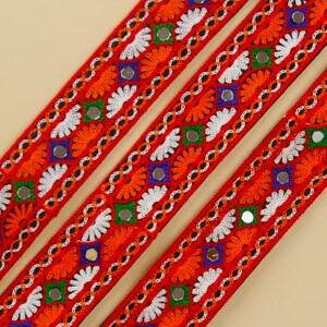 May include: Close-up of three red embroidered ribbons with intricate patterns. The ribbons feature orange, white, green, and blue floral designs, accented with small mirrored embellishments. The ribbons are diagonally arranged against a neutral background.