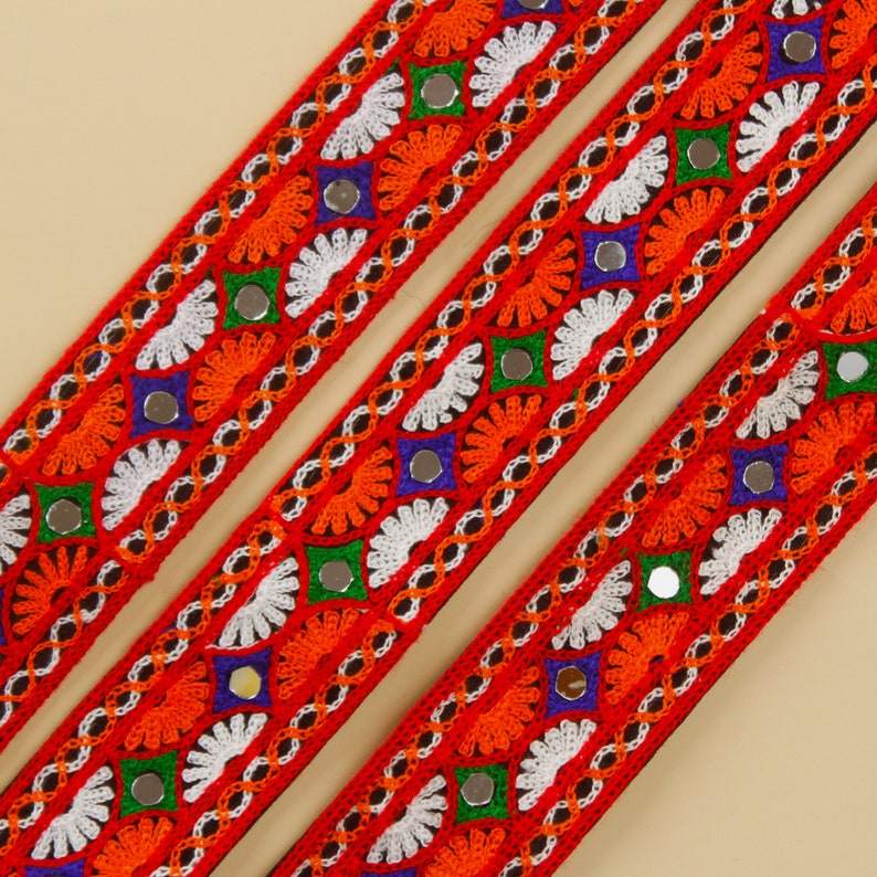 May include: Close-up of three red embroidered ribbons with intricate patterns. The ribbons feature orange, white, green, and blue floral designs, accented with small mirrored embellishments. The ribbons are diagonally arranged against a neutral background.