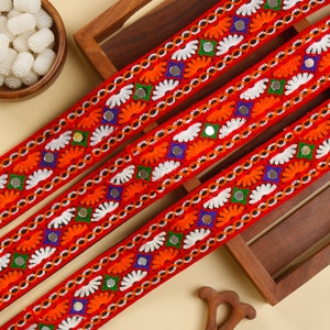 May include: Three vibrant red embroidered ribbons with intricate patterns. The design includes orange, white, green, and blue floral and geometric elements, along with small reflective mirror accents. The ribbons are displayed on a wooden surface.