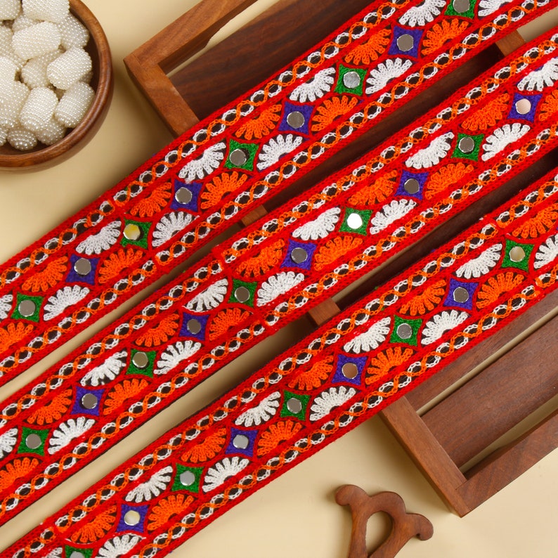 May include: Three vibrant red embroidered ribbons with intricate patterns. The design includes orange, white, green, and blue floral and geometric elements, along with small reflective mirror accents. The ribbons are displayed on a wooden surface.