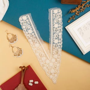 Könnte beinhalten: Elfenbeinfarbener Spitzenkragen mit floralen Stickereien und Perlenakzenten. Goldfarbene Creolen mit perlenartigen Perlen werden neben dem Kragen präsentiert. Die Artikel sind auf einer türkis- und beigefarbenen Oberfläche angeordnet.