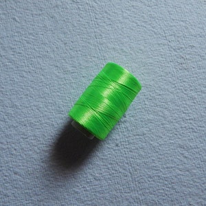 May include: A bright green spool of thread, tightly wound, sits on a light blue textured surface. The thread has a shiny, almost satin-like appearance, suggesting it may be silk or a similar material. The image is a close-up, highlighting the thread's vibrant color.