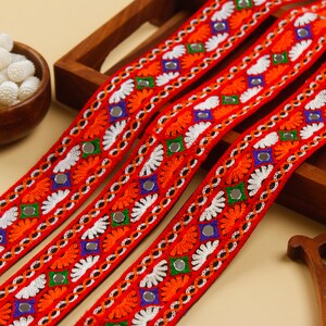 May include: Four strands of vibrant red embroidered trim with intricate floral and geometric patterns. The trim features orange, white, green, and blue accents, along with small mirrored embellishments. The trim is likely used for decorative purposes.