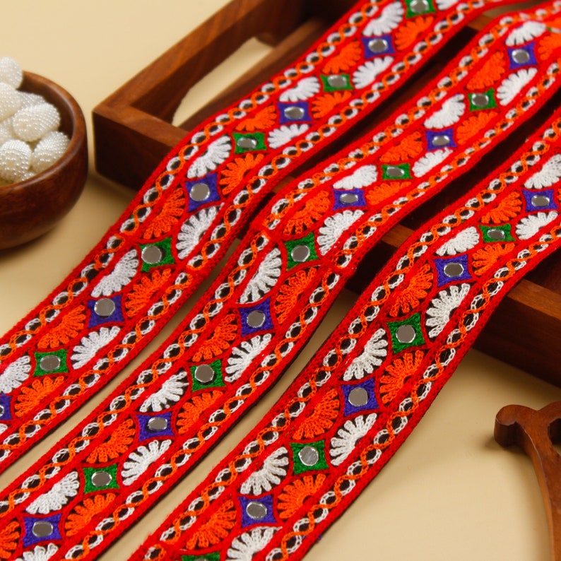 May include: Four strands of vibrant red embroidered trim with intricate floral and geometric patterns. The trim features orange, white, green, and blue accents, along with small mirrored embellishments. The trim is likely used for decorative purposes.