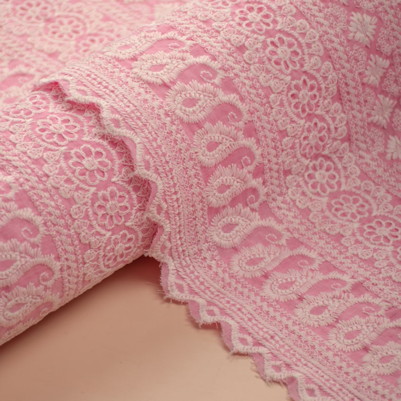 May include: Pink fabric with white embroidered paisley and floral designs. The fabric is rolled up and the edges are scalloped.
