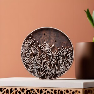 May include: A round wooden block with a carved design of flowers and leaves. The block is brown and has a natural wood finish.