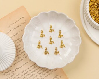 May include: A collection of small, gold-colored charms in the shape of a seated figure, arranged on a white, scalloped dish. The charms are likely for jewelry making or crafting. The background includes a book and a decorative dish.