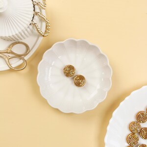 May include: Two gold-coloured, circular earrings with a textured, spiral design rest on a white, scalloped-edge dish. The earrings are on a light yellow surface. Other white dishes and gold accessories are also visible.