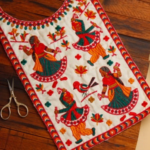May include: White fabric with a colorful embroidered design featuring four figures dancing. The figures are dressed in red, green, and yellow clothing. The fabric has a decorative border with red, orange, and yellow zigzags.