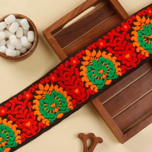 May include: Black embroidered trim with red, orange and green floral designs. The trim is displayed on a wooden tray.