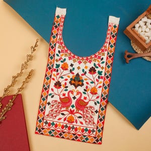 Puede incluir: Textil bordado de color crema con cuello en forma de U. El diseño presenta dos pavos reales enfrentados, motivos florales y un borde de formas geométricas en rojo, naranja, verde y amarillo. El textil es probablemente una pieza decorativa o parte de una prenda.