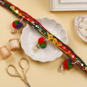 以下が含まれることがあります： 黒と緑のトリムにカラフルなポンポンとミラーのアクセントが施されています。トリムは白いプレートの上にあり、糸巻きが2つとハサミが1つあります。