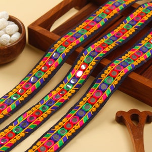 May include: A close-up of three navy blue fabric trims with colourful embroidery and round mirror accents. The trim features pink, green, blue, and yellow thread.