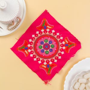 Puede incluir: Un cuadrado de tela rosa intenso con intrincados bordados. El diseño presenta un patrón circular central con detalles azules, verdes y blancos, rodeado de motivos florales estilizados en naranja, verde y azul. Los bordes de la tela están deshilachados.