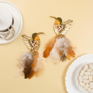 Könnte beinhalten: Ein Paar goldene Kolibri-Ohrringe mit braunen, weißen und beigefarbenen Federn. Die Kolibris sind mit Pailletten, Perlen und Perlen verziert.
