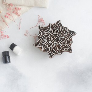 May include: A wooden block printing stamp with a detailed floral design. The stamp is brown with black lines and dots. It is sitting on a white surface with two small bottles of liquid.