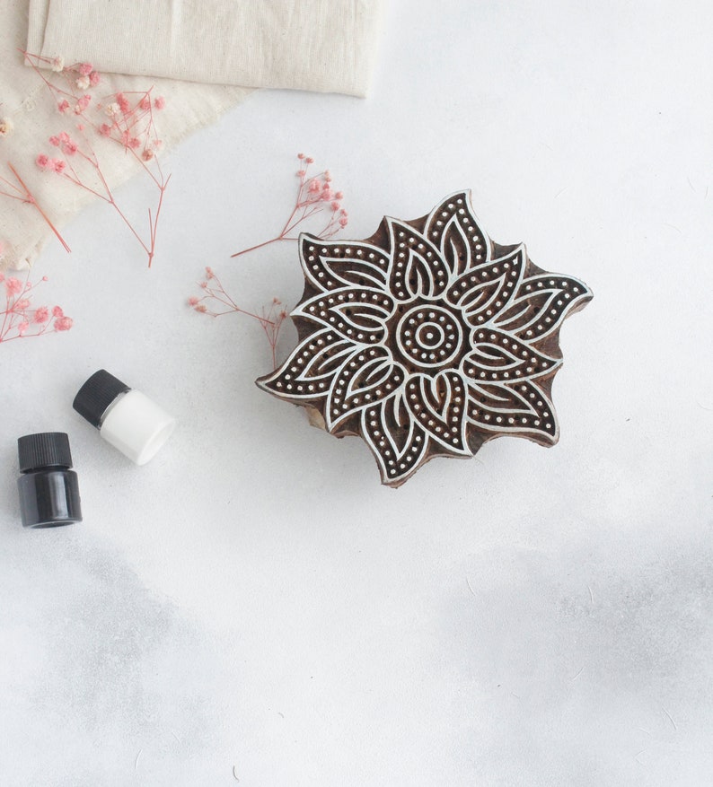 May include: A wooden block printing stamp with a detailed floral design. The stamp is brown with black lines and dots. It is sitting on a white surface with two small bottles of liquid.