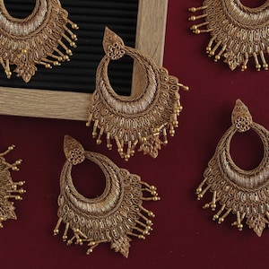 Op de afbeelding: Vijf gouden hangende oorbellen met ingewikkelde ontwerpen en kralenranden. De oorbellen zijn gerangschikt op een bordeauxrode ondergrond.