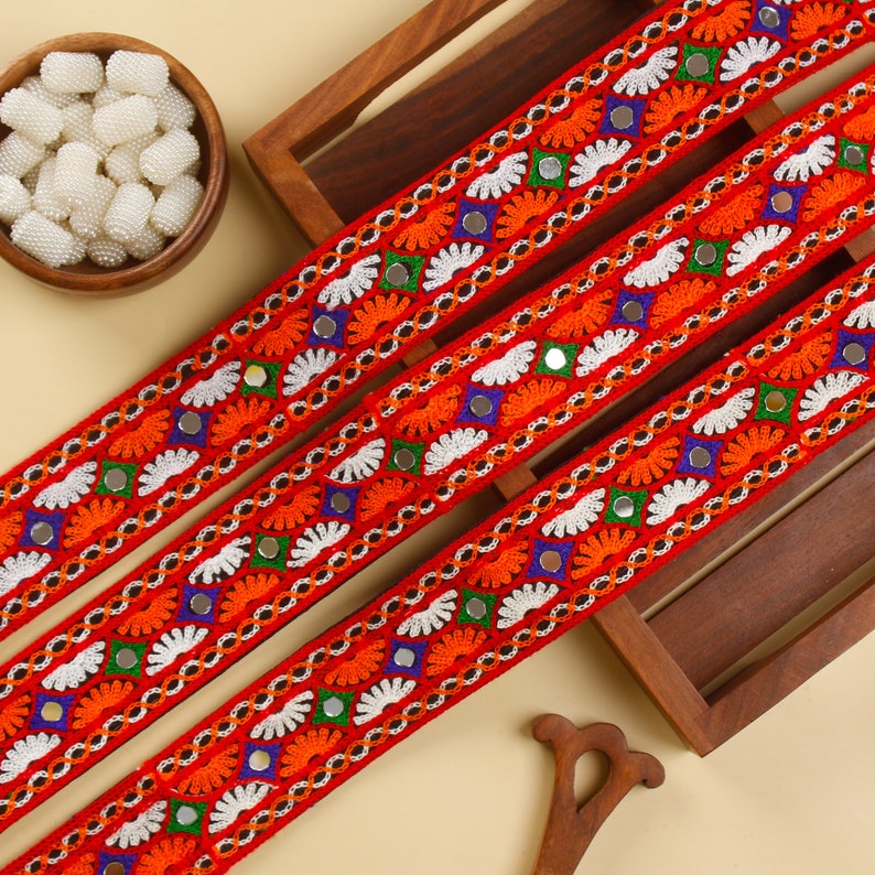 May include: Close-up of three red embroidered ribbons with intricate patterns. The ribbons feature white, orange, blue, and green floral designs, along with small mirrored accents. A wooden bowl of white beads and a wooden tray are in the background.