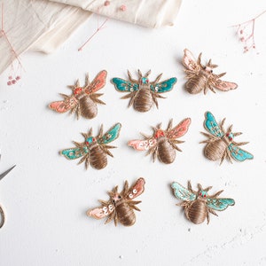 Può includere: Otto toppe ricamate a forma di ape in vari colori, tra cui oro, verde acqua e rosa. Le toppe sono disposte in cerchio su una superficie bianca.