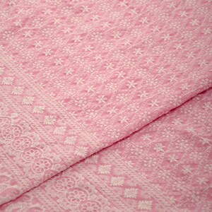 May include: Pink fabric with white embroidered floral patterns. The fabric has a delicate, intricate design.