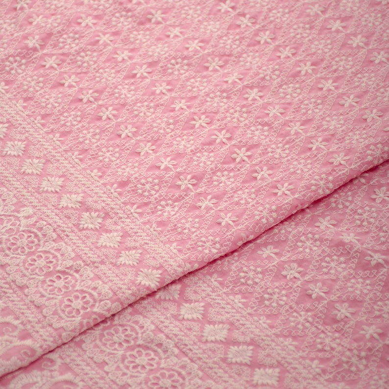 May include: Pink fabric with white embroidered floral patterns. The fabric has a delicate, intricate design.