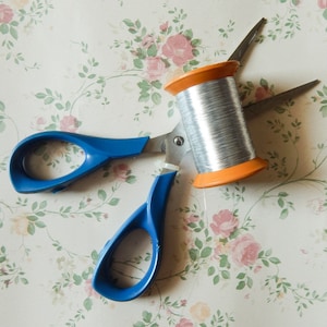 May include: A pair of blue-handled scissors with silver blades, positioned near a spool of silver thread with an orange core. The items are set against a floral patterned background with pink and peach roses.