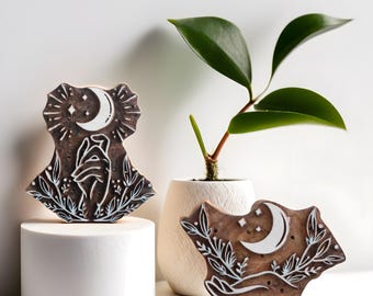 Puede incluir: Dos bloques de impresión de madera con un diseño de luna creciente y estrellas. Los bloques son marrones con detalles blancos y presentan una mano que alcanza la luna. Los bloques se muestran sobre pedestales blancos sobre un fondo blanco.