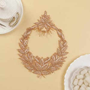 Può includere: Applicazione ricamata dorata e dettagliata a forma di ghirlanda, con intricati disegni di foglie e fiori. L'applicazione è impreziosita da piccole perline e paillettes, perfetta per abbellire capi d'abbigliamento o lavori artigianali.