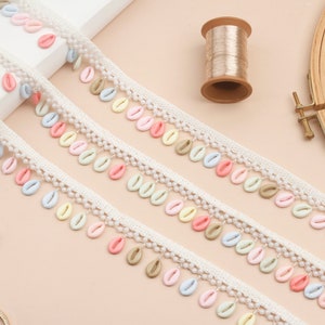 以下が含まれることがあります： パステルカラーの貝殻型装飾が施されたクリーム色のレーストリム。トリムには、小さな丸いビーズと、ピンク、青、黄色、緑のカラフルな貝殻のアクセントが付いています。糸巻き、ハサミ、刺繍枠も写っています。