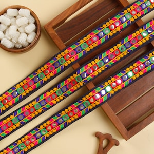 May include: Three strips of black fabric trim with colorful embroidery and round silver mirrors. The trim is displayed on a wooden tray.