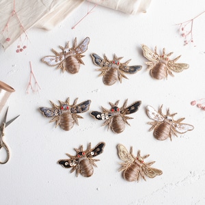Peut inclure: Huit patchs brodés en forme d'abeilles dans différentes couleurs, dont l'or, le blanc, le noir et le bleu. Les abeilles ont des yeux en perles rouges et sont disposées sur une surface blanche.
