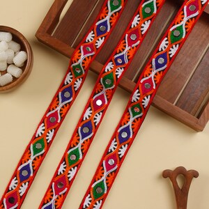 May include: Three strands of colorful embroidered trim with mirror accents. The trim is red, orange, green, blue, and white. The trim is on a wooden surface.