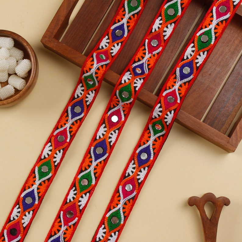 May include: Three strands of colorful embroidered trim with mirror accents. The trim is red, orange, green, blue, and white. The trim is on a wooden surface.