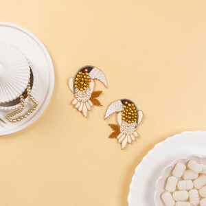 May include: A pair of gold and white toucan earrings with black eyes and beaks. The earrings are made of beads and are on a light yellow background.