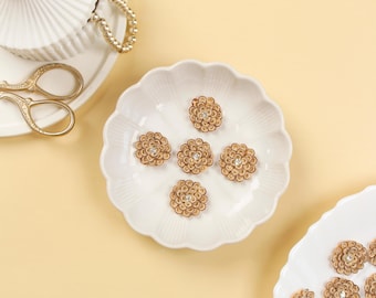 May include: A top-down shot shows a white scalloped dish holding six brown flower-shaped embellishments with a central rhinestone. Other white dishes, gold scissors, and jewelry are also visible on a light yellow surface.